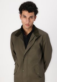 Olive green coat with a corduroy collar, snap buttons, and side pockets, displayed on a model against a white background.
