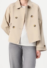 Beige värinen lyhyt takki, joka on valmistettu pehmeästä kankaasta. Siinä on suuret ruskeat napit, leveä kaulus ja sivutaskut. Päällä valkoinen paita.