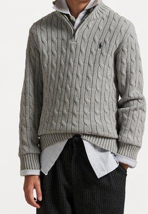 Person iført grå kabelstrikket sweater med kvart lynlås over blå stribet skjorte med knapper og sorte bukser med nålestriber, hænder afslappet langs siderne.