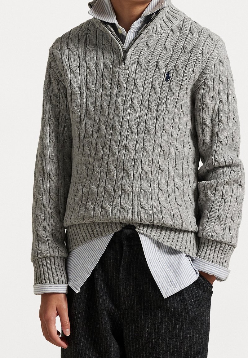 Personne portant un pull gris en maille torsadée avec fermeture éclair jusqu'à mi-poitrine sur une chemise bleue à rayures boutonnée et un pantalon noir à fines rayures, mains détendues sur les côtés.