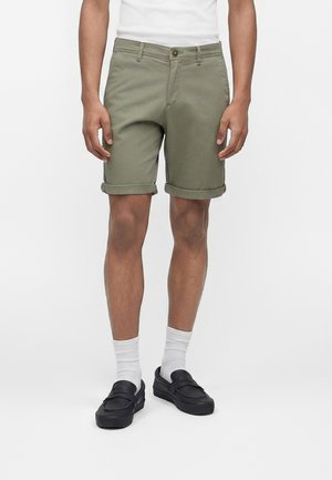 Bărbat purtând pantaloni scurți verde măsliniu suflecați, șosete albe până la jumătatea gambei, mocasini negri și o cămașă albă, stând în fața unui fundal simplu.