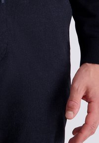 Donkerblauwe trui met een getextureerde stof, voorzien van een geribde boord en een ritsdetail aan één kant, gedeeltelijk bedekt door een hand.