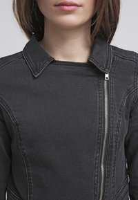 Veste en denim noire avec un col pointu, une coupe ajustée et un détail de fermeture éclair argentée sur le devant, présentant un tissu texturé.