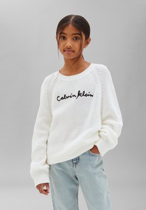 Jeune fille aux cheveux foncés portant un pull en maille blanc Calvin Klein et un jean bleu clair, debout devant un fond gris uni.