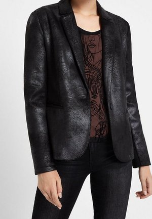 Person wears a textured black blazer over a sheer black top with line art faces, paired with black skinny jeans against a plain background.