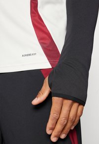 Long-sleeve athletic top in black and white with red accents. Features a stretchy, moisture-wicking fabric with thumbhole cuffs and logo detail.