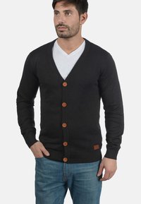Mörkgrå stickad cardigan med v-ringning, utrustad med sju bruna knappar, ribbad manschett och en texturerad finish, tillsammans med en vit t-shirt under.