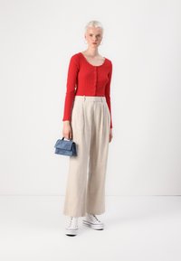Haut à manches longues rouge à côtes avec détails de boutons, associé à un pantalon large beige et des baskets à plateforme blanches. Un petit sac à main bleu vient parfaire le look.