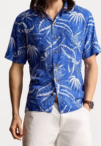 Niebieska koszula z krótkim rękawem z białym wzorem tropikalnych liści, z drewnianymi guzikami i luźnym krojem. Nosi się ją z jasnymi spodniami.