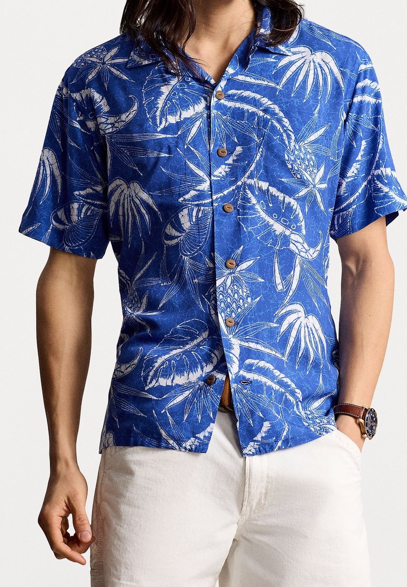 Niebieska koszula z krótkim rękawem z białym wzorem tropikalnych liści, z drewnianymi guzikami i luźnym krojem. Nosi się ją z jasnymi spodniami.