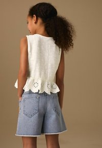Fille aux cheveux bouclés portant un top blanc sans manches avec un bord ajouré et un short en jean bleu clair, debout devant un fond beige.