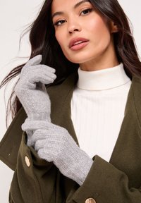 Hallid kootud sõrmikud tekstuurse mustri ja soonilise disainiga, mida kannab modell valge kõrge kaeluse ja oliivrohelise mantliga.