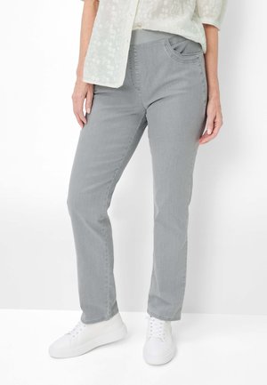 Kvinde iført lysegrå jeans med lige ben, hvide sneakers og en hvid broderet bluse med korte ærmer, stående mod en hvid baggrund.