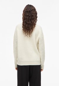 Άτομο με μακριά καστανά σγουρά μαλλιά που φοράει ένα κρεμ πλεκτό πουλόβερ και μαύρο παντελόνι, γυρισμένο προς τα πίσω απέναντι από ένα λευκό φόντο.