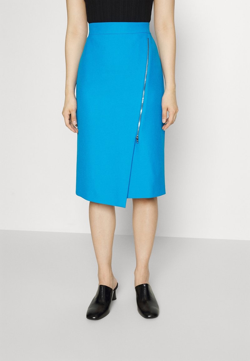 Femme portant une jupe bleu vif longueur genou avec une fermeture éclair diagonale à l'avant, un haut noir et des chaussures noires sans lacets, debout sur un sol gris.