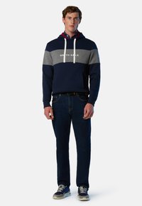 Sweatshirt navy et gris avec une poche frontale, capuche avec cordon de serrage, et logo "NORTH SAILS" sur la poitrine, associé à un jean foncé et des baskets.