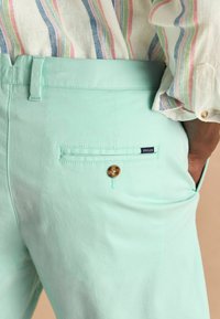 Minti zaļas bikses ar aizmugurējo kabatu, kurā ir zelta pogas, ar zīmola etiķeti "Joules" un gludu kokvilnas auduma tekstūru.
