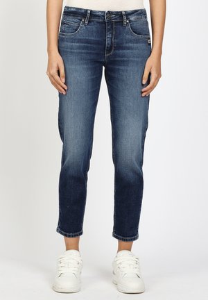 Person trägt dunkelblaue, eng anliegende Jeans und weiße Turnschuhe, steht vor einem schlichten hellen Hintergrund.