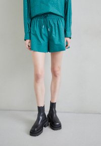 Pantalones cortos en color azul verdoso con cintura elástica y cordón, combinados con botas de cuero negras hasta el tobillo que presentan suelas gruesas y costuras decorativas.