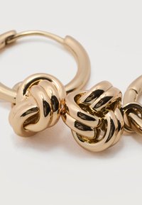 Κοντινό πλάνο λαμπερών χρυσών σκουλαρικιών κρίκων με σχέδιο δεσμού κόμπου, σε λευκό φόντο.