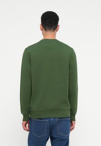 Férfi háttal áll, sima zöld környakas pulóverben és kék farmerben, egy fehér háttér előtt.