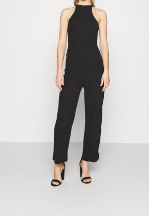 Kobieta ubrana w czarny kombinezon bez rękawów z wysokim dekoltem oraz czarne sandały na wysokim obcasie, stojąca na jednolitym tle.