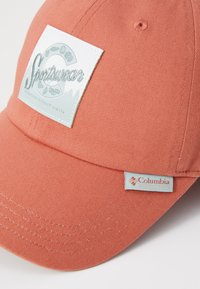 Korall baseballcap med et hvitt logo-patch som viser "Sportswear" og fjellgrafikk; inkluderer et Columbia-merkelapp på siden.