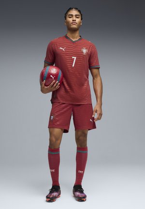 Calciatore maschio che indossa la divisa rossa del Portogallo e tiene in mano un pallone da calcio rosso abbinato, in piedi su uno sfondo grigio uniforme.