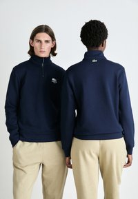 Pulover s visokim ovratnikom u tamnoplavoj boji sa zipom i Lacoste logotipom na prsima. Mekani materijal s rebrastim manžetama i rubovima.
