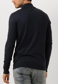 Mausblauwe pullovertrui met een hoge hals en lange mouwen, met een soepele textuur en ribgebreide manchetten, gecombineerd met lichtgrijze jeans.