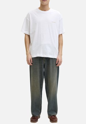 Person iført løs hvid crewneck T-shirt med lille tekst på brystet, brede falmede grå jeans og brune lædersko, stående med front mod kameraet.