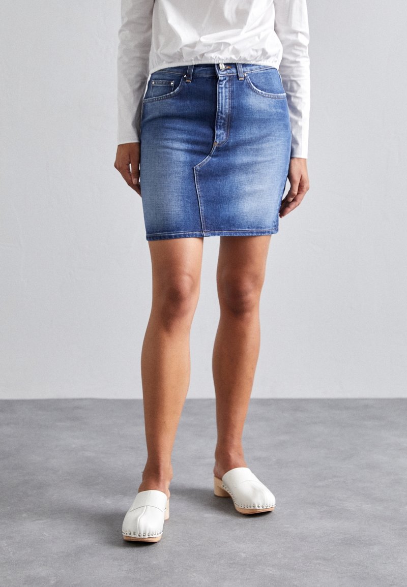 Femme portant une mini-jupe en jean bleu délavé, une chemise blanche à manches longues et des sabots blancs sans bride, debout sur un sol gris.