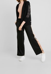 Sweat à capuche noir zippé avec poignets côtelés et texte logo sur les manches, associé à un pantalon large noir avec des bandes latérales pêche et des boutons-pression.