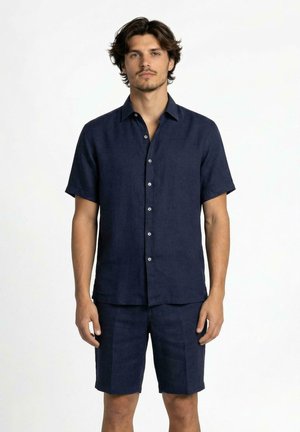 Giovane uomo con capelli castani spettinati che indossa una camicia blu navy a maniche corte con bottoni e pantaloncini blu navy abbinati, in piedi su sfondo bianco.