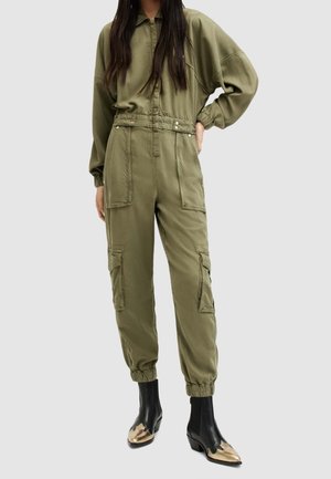Woman wearing olive green utility jumpsuit with front pockets and black ankle boots featuring metallic gold toe caps.