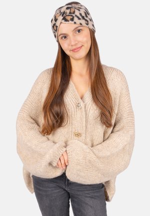 Beige Strickjacke mit übergroßen Ärmeln und zwei goldenen Knöpfen. Kombiniert mit einem Kopfband im Leopardenmuster in warmen Farbtönen. Dunkle Jeans.