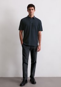 Ciemnoniebieska koszulka polo z trzema guzikami, noszona z dopasowanymi czarnymi spodniami i czarnymi butami wiązanymi, stojąca na neutralnym tle.