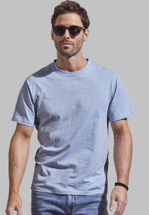 Helles blaues T-Shirt mit Rundhalsausschnitt aus weichem Stoff, kurze Ärmel, kombiniert mit dunklen Sonnenbrillen und einem Perlenarmband.