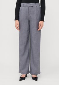 Grijze stoffen wijde broek met voorplooien, een tailleband en zijzakken. Gedragen met een zwart topje en zwarte hoge hakken.