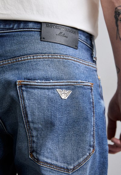 Jeans din denim de culoare albastră medie, cu o etichetă din piele pe spate inscripționată "Emporio Armani Milano" și un logo argintiu cu un vultur pe buzunar.