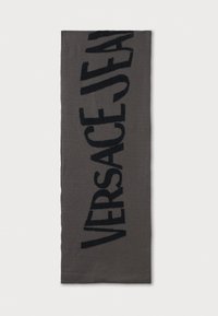 Sivi šal s mekom teksturom, s istaknutim crnim logoom "VERSACE JEANS" u debelim slovima. Pravokutnog oblika, minimalističkog dizajna.