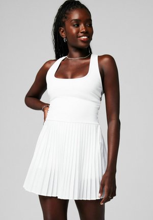 Frau mit geflochtenem Haar, die ein ärmelloses weißes Kleid mit quadratischem Ausschnitt und plissiertem Rock trägt, lächelt und steht vor einem schlichten Hintergrund.