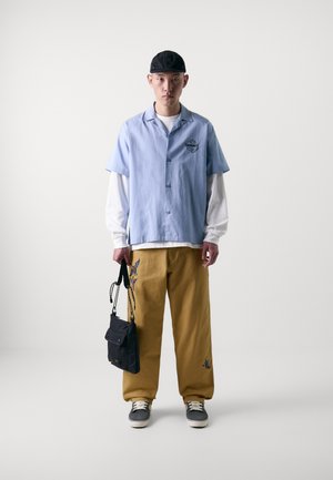 Himmelblaues Kurzarmhemd, darunter ein weißes Langarmhemd, gelbe Hose mit Stickerei, schwarze Tasche und graue Schuhe mit weißen Sohlen.