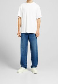 T-shirt bianco a maniche corte abbinato a jeans in denim blu chiaro a gamba larga e sneakers bianche, in piedi contro uno sfondo grigio uniforme.