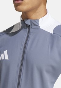 Grijze sportjack met een hoge kraag, witte accenten op de schouders, een ritssluiting aan de voorkant en een logo op de borst. Glad, ademend materiaal.
