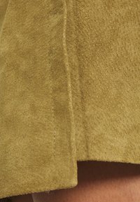 Tessuto scamosciato verde oliva con una superficie morbida e testurizzata. Impunture visibili lungo l'orlo, che evidenziano la forma e le linee dell'indumento.