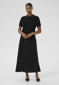 Robe noire avec un haut en tricot à manches courtes et un col à rabat, agrémenté de détails en boutons, associé à une jupe longue et fluide avec une fente à l'avant.
