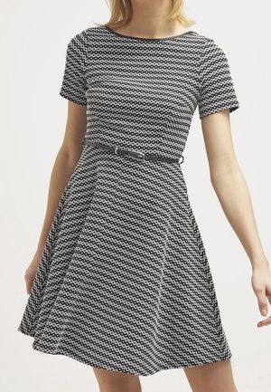 Vrouw draagt een zwart-witte jurk met korte mouwen en een dunne zwarte riem in de taille.