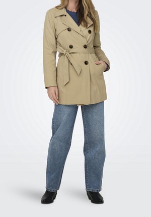 Donna che indossa un trench beige a doppio petto sopra un maglione blu, con jeans blu e stivaletti neri, in posa con una mano in tasca.