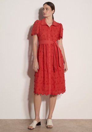 Kobieta stojąca w czerwonej sukience z krótkim rękawem, sięgającej do kolan, ozdobionej kwiatową haftem, z guziczkami z przodu, kołnierzykiem, paskiem w talii oraz beżowymi, niskimi sandałami z odkrytymi palcami.